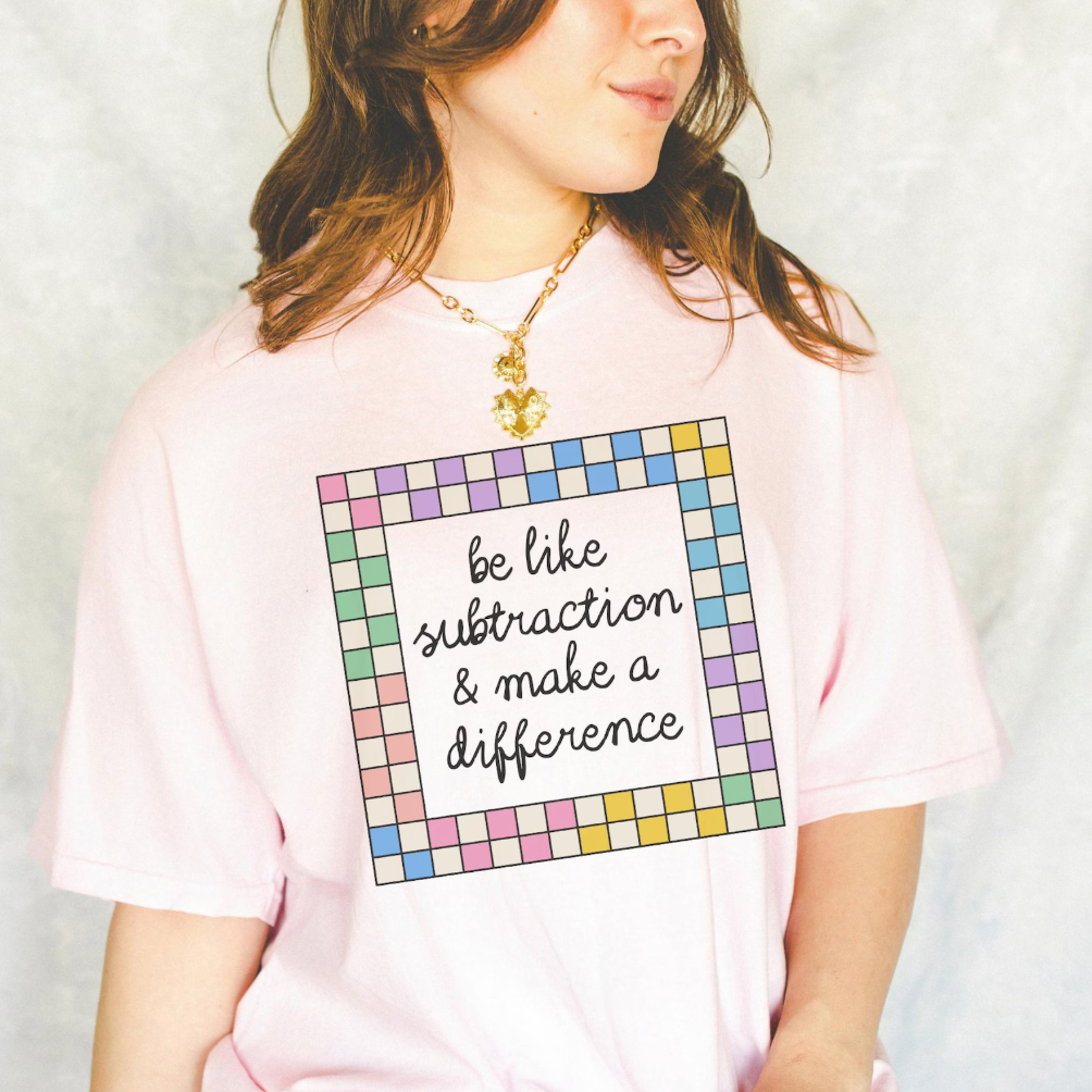 A woman wearing a light pink t-shirt with a colorful grid-patterned graphic that reads 'be like subtraction & make a difference' in black script. She is wearing blue jeans and a gold necklace, posing against a light gray textured backdrop.