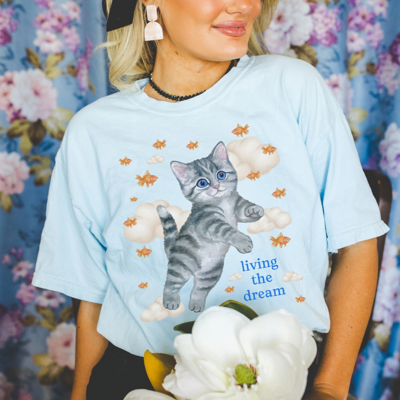 Person wearing a light blue t-shirt with a cat graphic and text, standing against a floral curtain background.