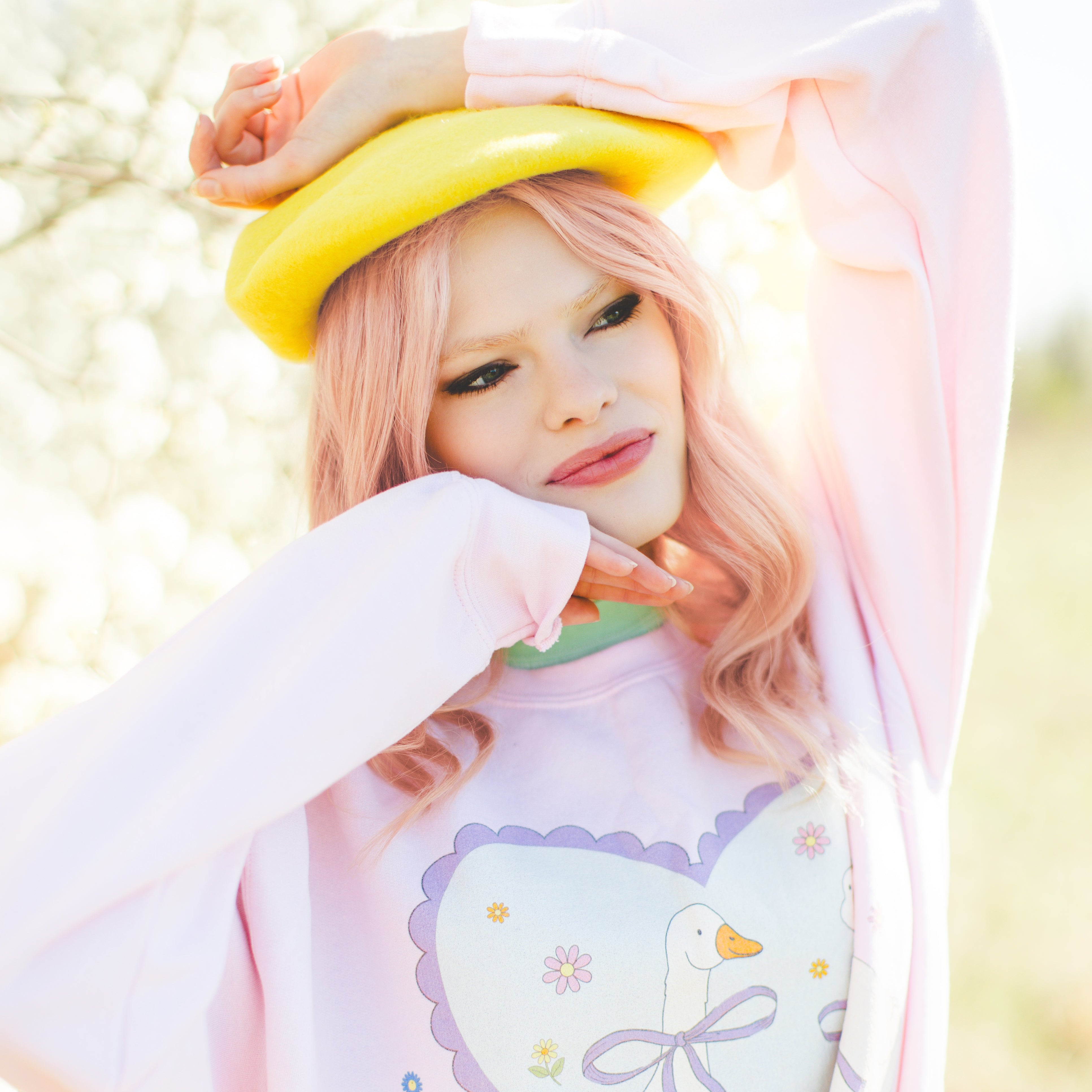 Pink haired woman wearing a pastel-colored cute goose sweatshirt and a yellow hat, standing outdoors.