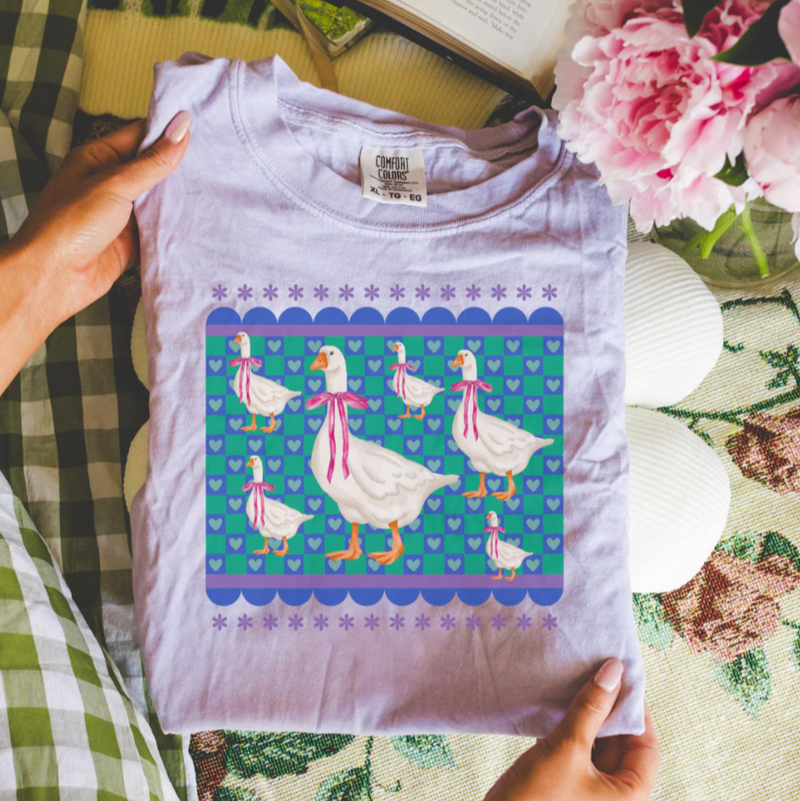 purple t-shirt with colorful duck design held by a person on a checkered fabric background with flowers.