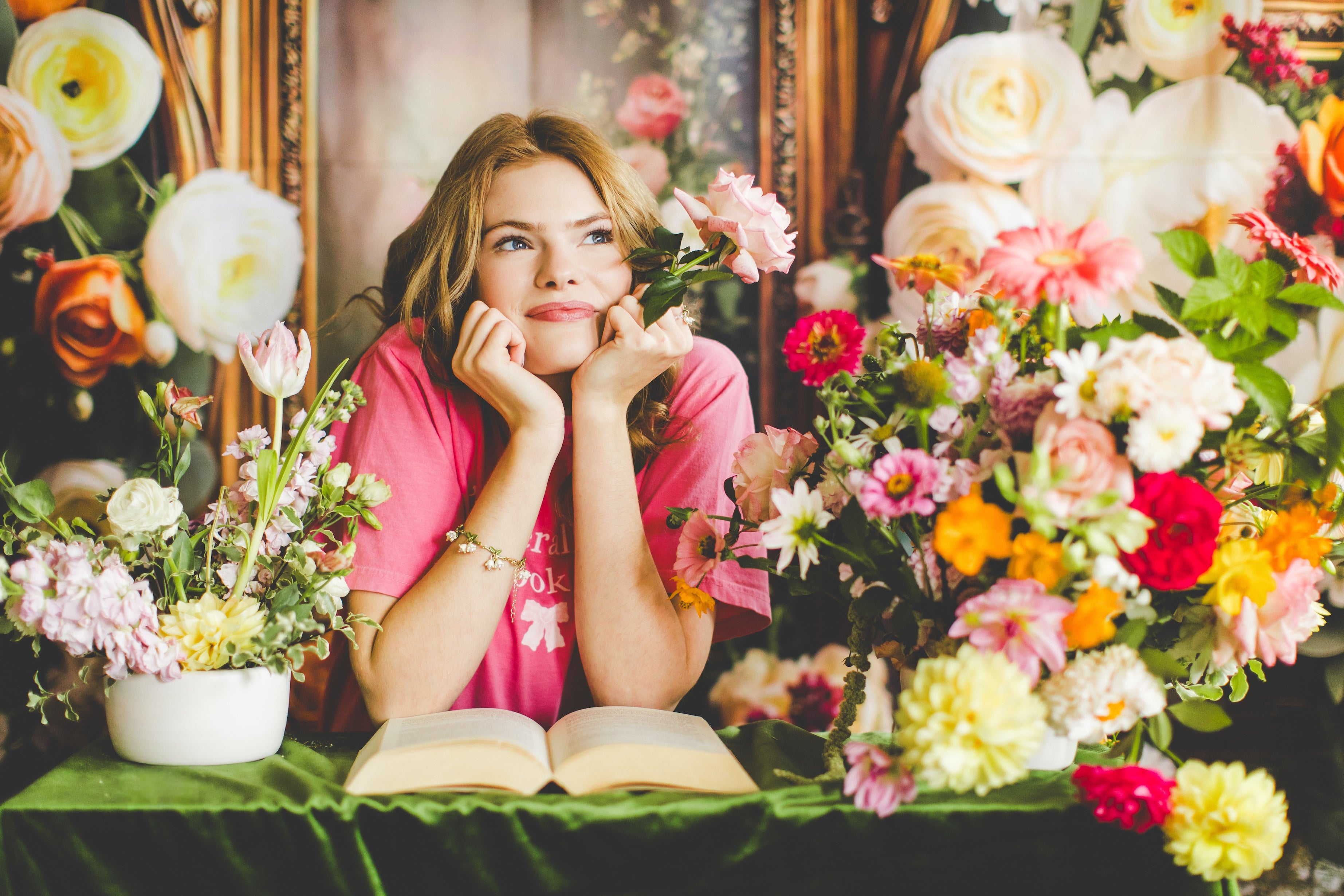 Woman wearing a bookish shirt sitting among flowers with an open book, surrounded by vibrant floral arrangements.